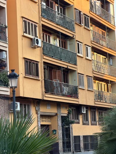 The image shows a multi-story apartment building in Benimàmet-beniferri, Spain. The building has an orange-yellow facade with several balconies featuring black metal railings. The windows are fitted with brown wooden shutters, some of which are open, revealing interior details. Air conditioning units are visible mounted on the exterior walls below some of the windows. A black vintage-style street lamp stands in the foreground to the left, partially obscured by lush green palm fronds. The scene is illuminated by warm sunlight, suggesting it was taken during the late afternoon or early morning. The building appears to be in a residential area.