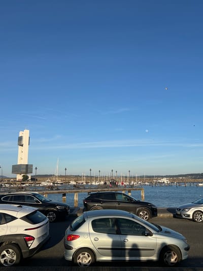 The image depicts a marina scene in La Coruña, Spain, characterized by a clear blue sky with minimal cloud cover. The white "Torre de Control Marítimo de A Coruña" is prominent on the left, a multi-story, angular white structure with dark glass panels. A bright, nearly full moon is visible in the upper center-right sky, and several small birds are in flight.
In the foreground, four vehicles are parked along what appears to be a paved waterfront area. From left to right: a white compact SUV with black trim, a black SUV, and a silver two-door hatchback are clearly visible. The front portion of a silver sedan is partially visible on the far right. Shadows from the cars and unseen foreground objects extend towards the lower right, indicating a high sun position.
Beyond the parked cars, a body of water—the marina—is populated by numerous sailboats and powerboats docked at piers. A long wooden or concrete pier with multiple streetlights extends across the mid-ground, supporting various parked bicycles and a few small groups of individuals walking or standing. The background features distant landforms, likely hills, under the bright sky. No specific interactions between people are clearly discernible, but their presence suggests recreational activity. The overall impression is one of a clear, bright day at a busy port.