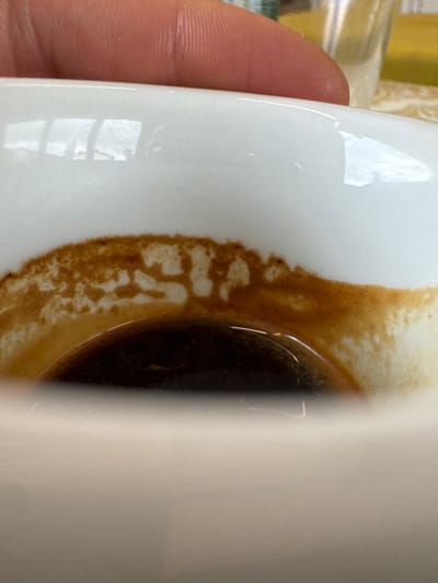 The image presents a close-up, downward-angled view into a white ceramic cup, located in Anzola dell'Emilia, Italy. The cup contains a small volume of dark brown liquid, consistent with coffee, at its base. The interior wall of the cup, above the liquid line, is marked by a significant deposit of dried, reddish-brown granular residue, likely coffee crema, forming an irregular, textured band. Within this residue, distinct shapes resembling the letters "SI" or the digits "51" are discernible. A human hand, specifically a portion of the thumb and possibly an index finger, is visible at the top of the frame, partially obscuring the cup's upper rim. Reflections on the polished surface of the cup indicate an indoor environment with overhead light sources or windows. The background above the cup is blurred, revealing indistinct shapes and a pale yellow surface.
