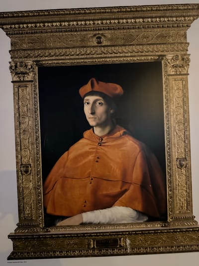 The image shows a framed portrait painting of a man wearing an orange cap and robe. The man has light skin and is looking to his left. The background of the painting is dark. The frame is ornate and gold-colored. Below the frame, there is a label that reads "RAFAEL". At the very bottom of the image, a small text indicates "Museo Nacional del Prado - 2019". The location of Puerto de Mazarrón, Spain is not indicated in the image.