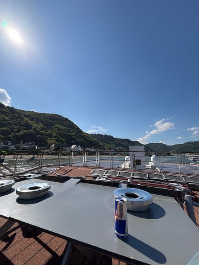 The image shows a view from a boat or ship's deck, overlooking the Rhine River and the town of Kestert, Germany. The foreground features a gray surface with two circular metal objects, possibly covers for access points, and a Red Bull can standing upright with its top open. The middle ground shows a metal railing and a body of water with ripples reflecting the sunlight. In the background, the town of Kestert is visible, nestled at the foot of a green, tree-covered hill. Several houses with dark roofs are situated along the riverbank. The sky is clear blue with a bright sun in the upper left corner, casting a lens flare. The scene depicts a sunny day on the Rhine River, with the Red Bull can suggesting a moment of refreshment during the journey.