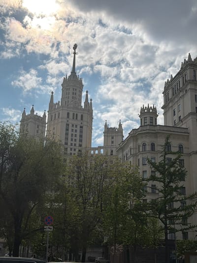 The image shows a tall, ornate building with a spire against a cloudy sky. The building is light-colored and features multiple tiers with decorative architectural details. Trees with green leaves partially obscure the building at the lower portion of the frame. A traffic sign with a red and blue "X" is visible on the right side of the image, partially hidden by tree branches. The sun is behind the clouds on the left side of the image, creating a bright glow. The image appears to be taken from a low angle, looking up at the building and sky. I cannot make claims about the location beyond what is provided in the image.
