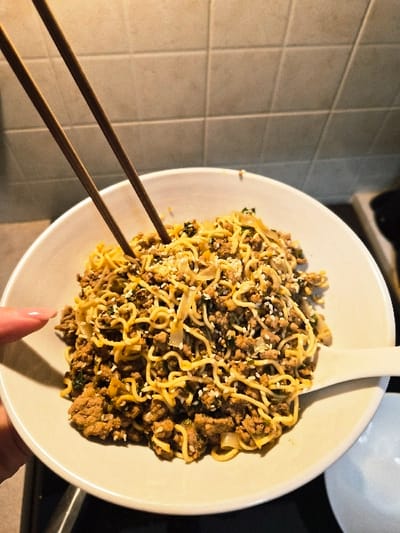 The image depicts a close-up view of a bowl of food, held by a person's hand, within an indoor setting. The specific location within the Municipal Unit of Corinth, Greece, cannot be determined from the visual information provided.

The primary subject is a large, light-colored ceramic bowl filled with a meal composed of yellow curly noodles, brown granular ground meat, green leafy components, and white sesame seeds sprinkled throughout. Two brown chopsticks are inserted vertically into the center of the meal. A white ceramic spoon rests on the right rim of the bowl. A human hand, with a pink-painted fingernail on the index finger, is visible on the lower left, gripping the exterior of the bowl.

The background features a light-colored tiled wall, likely a kitchen backsplash, with visible grout lines forming a grid pattern. Below the bowl, on the left, a dark, indistinct surface is present. On the lower right, a portion of a smaller, white ceramic dish or bowl is visible. Illumination appears to be artificial, casting some subtle shadows.