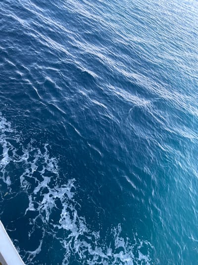 The image displays a close-up, high-angle view of the sea's surface. The water transitions from a deep blue in the lower left to a lighter, almost turquoise hue towards the upper right, indicating shallowing or a change in depth. Numerous small ripples and waves are visible across the entire surface, reflecting the ambient light and creating a textured pattern. In the lower left quadrant, white foam and bubbles are present, suggesting the wake of a moving vessel. A portion of a white railing or boat edge is visible in the extreme lower left corner, running diagonally upwards. The scene is devoid of any people or specific landmarks, offering a purely abstract composition of water and light. Based on the provided location of Municipal Unit of Kastro - Kyllini, Greece, this image likely depicts a view of the Ionian Sea from a boat or coastline in that region.