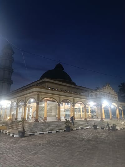 The image shows a mosque in Bugakkrueng, Indonesia, at night. The mosque has a dome and a minaret to the left. The front of the mosque has a series of arches supported by columns. There are several potted plants in front of the building. A man is walking up the stairs to the mosque's entrance. A bicycle is parked on the paved area in front of the mosque. The sky is dark blue, suggesting it is either dusk or night. The mosque is well-lit, contrasting with the dark sky.