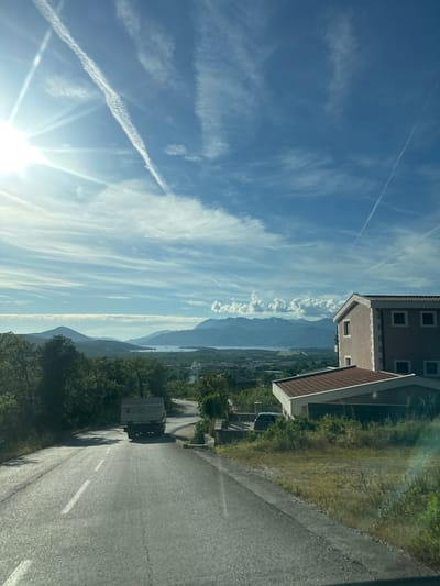 The image captures a panoramic daytime view in Pelinovo, Montenegro. The sky is clear blue with scattered white cirrus clouds and multiple linear aircraft contrails; one prominent contrail exhibits dispersion and segmentation across the upper left and central sky. A bright sunburst is positioned in the upper left quadrant, casting lens flare.
The mid-ground and background feature a mountainous landscape, with several layers of dark green and grey mountains extending to the horizon. Between these mountains, a large body of water, likely a bay or a fjord, is visible. Adjacent to the water in the mid-ground, a flat, rectangular area consistent with an airport runway is discernible.
In the foreground, a two-lane asphalt road with a dashed white center line slopes upwards and curves gently to the right. A light grey dump truck is observed driving up this road, its rear visible. To the right of the road, a two-story building with a light beige facade, white corner quoins, and a red tiled gabled roof stands. Below and slightly in front of it, a single-story structure with a similar red tiled roof is partially visible. A dark-colored vehicle is parked on a property behind the lower building. The roadside and surrounding areas are covered with green trees, bushes, and overgrown grass.