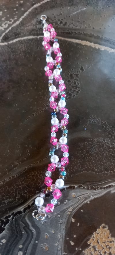 The image shows a close-up, overhead view of a beaded bracelet lying on a dark, textured surface. The bracelet is made of alternating pink faceted beads, white pearl beads, and smaller clear and iridescent beads. The bracelet is positioned vertically and appears to be laid out on a table or countertop with a swirling, abstract pattern. There are no people or specific landmarks of Iskra, Bulgaria visible in the image. The lighting is dim, with reflections on the surface suggesting it may be glossy.