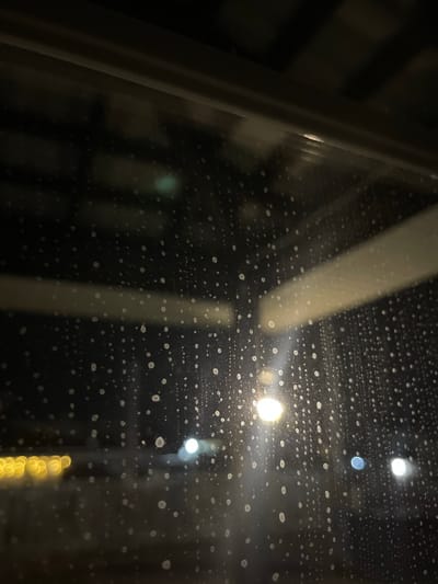 A low-angle, close-up view through a wet window, obscured by numerous water droplets. Outside, blurry lights are visible, including what appear to be streetlights and possibly string lights. The surroundings are dark, suggesting nighttime. A window frame is visible on the left side of the image. The location is unspecified and cannot be discerned from the image. There is no information available about people, subjects, objects, actions, or interactions other than what is stated.