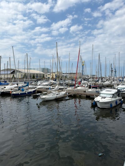 The image depicts a harbor in Wismar, Germany, filled with numerous sailboats and a few motorboats docked. The sailboats have tall masts, some with sails furled, others without. A prominent white motorboat with the name "Pajarito" is in the foreground, facing right. In the background, industrial buildings and cranes are visible, suggesting a port or shipbuilding area. The water is dark and reflective, with ripples creating distorted reflections of the boats and sky. The sky is partly cloudy with blue patches. There are no people visible in the image.