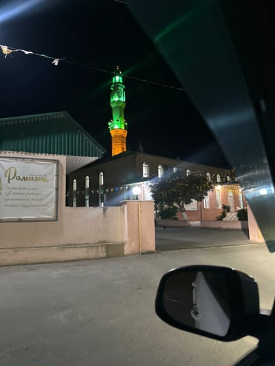 This is a vertical, nighttime photograph taken from inside a vehicle. The scene depicts a street with buildings and a brightly lit minaret of a mosque, illuminated in green and orange. Trees decorated with festive lights are visible between the buildings. In the foreground, a side-view mirror reflects a portion of the vehicle's interior and the street.
A banner with text is displayed on the side of a building. The visible text, written in Cyrillic and possibly Arabic script, appears to be related to Ramadan. The text reads "Ramadan" in large golden script, with smaller text below.
The overall impression is one of a festive evening celebration, likely during the month of Ramadan, in a town with a prominent religious structure. The lighting suggests it is after sunset, and the presence of decorative lights implies a celebratory atmosphere. The reflection in the mirror adds a sense of perspective, indicating the image was captured while the viewer was in motion.