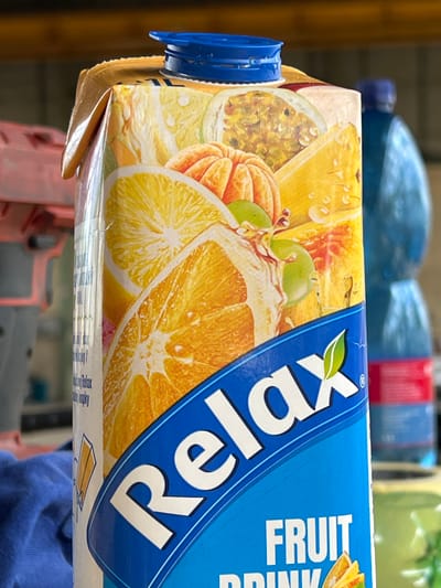 A carton of "Relax" brand fruit drink is shown, featuring images of various fruits like oranges, passionfruit, and grapes. The carton has a blue cap and a blue band across the bottom with the brand name. In the background, two large blue plastic bottles are visible. The scene appears to be indoors, possibly a warehouse or shop, given the presence of what looks like industrial equipment and shelves in the background. The lighting suggests a natural light source mixed with artificial light.
