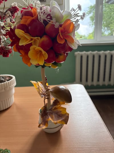 The image shows a close-up view of an artificial fruit and flower arrangement on a wooden table. The arrangement features a cluster of red and orange faux fruits, interspersed with yellow and orange artificial flowers and white decorative branches. Below the fruit cluster, the arrangement has a brown stem wrapped with copper wire, adorned with artificial autumn leaves. It is planted in a small, white pot. To the left of the arrangement, a white textured planter with soil and what appears to be a small plant is visible. In the background, a green wall is present, with a white radiator against it and a window showing trees and sky. The overall scene suggests an indoor setting, likely a room in a house or office in Ветрынскі сельскі Савет, Belarus, given the context. There are no people or animals visible in the image.