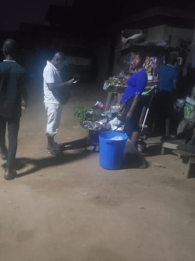 Here is a detailed description of the image:
**Location:** Alalubosa, Nigeria.
**Scene:** The image appears to be taken outdoors in what looks like a market or roadside stall environment at night.
**People:** Several people are visible, though visibility is limited.
* One person is wearing a blue shirt and appears to be interacting with the stall.
* Another person, closer to the camera, is wearing light-colored pants and a white shirt. They appear to be holding a phone or similar device.
* A third person, mostly in shadow, stands near the edge of the frame, wearing a dark jacket.
**Objects:** Key objects in the scene include:
* A roadside stall or display, stocked with various items.
* A blue plastic bucket sitting near the stall.
* Vegetables or produce on display.
**Actions/Interactions:**
* A person in the blue shirt appears to be shopping or inspecting items at the stall.
* The person in the white shirt is holding their phone and seemingly observing the scene.
**Environment:** The environment looks like a typical informal market area, likely at night. The ground is dirt or pavement. Lighting is poor.