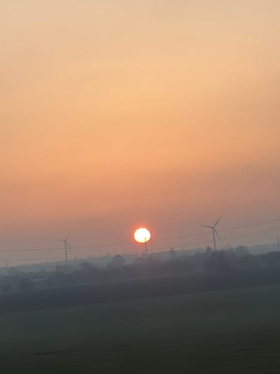 The scene depicts a rural landscape at either sunrise or sunset in Poruchik Chunchevo, Bulgaria. The upper two-thirds of the image feature a sky transitioning from a light yellow-orange at the top to a deeper orange near the horizon. A bright, circular, orange-yellow sun is positioned low in the sky, slightly left of the image's vertical center, partially obscured by a dark, thin vertical utility pole directly beneath it.

Horizontal utility lines stretch across the mid-ground, silhouetted against the sky, connecting to several indistinct poles. Two prominent, multi-bladed wind turbines are visible as dark silhouettes against the sky in the background; one is positioned to the left of the sun, and another further to the right. A significant layer of atmospheric haze or fog blankets the horizon, obscuring the lower portions of the turbines and creating a muted, blurred effect for distant elements. The lower third of the frame comprises a dark, flat expanse of land, consistent with a cultivated field, rendered in deep green-grey tones due to the low light. No individuals or active interactions are discernible within the frame.