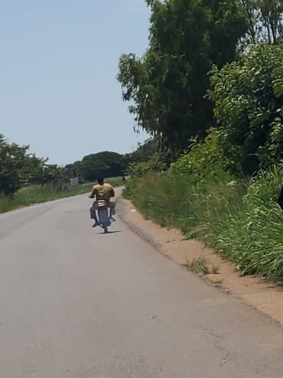 A person rides a motorcycle down a paved road in Mangu, Nigeria. The rider is wearing a yellow t-shirt and dark pants. The motorcycle is black. The road is gray and cracked, with a sandy shoulder on the right. Tall green grass lines the side of the road. In the background, there are trees and a clear blue sky. The image is taken from behind and to the right of the motorcycle.