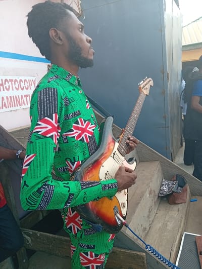 A young adult male, appearing Black, is depicted in profile facing left, holding and seemingly playing an electric guitar. He has dark, short hair and a trimmed beard. He wears a distinctive long-sleeved shirt and matching trousers, both green with a repeating pattern of white and red Union Jack flags interspersed with black text. The electric guitar is a distressed sunburst-finish instrument with a white pickguard and three single-coil pickups, featuring a blue coiled cable connected to its output jack. The subject is positioned on what appears to be a series of wooden steps or planks. To his immediate left is a light-colored wall displaying faded red text, partially legible as "PHOTOCOPY" and "LAMINATIO[N]". A large, dark rectangular structure stands behind him to his right. A second individual, wearing a blue shirt, is partially visible behind this structure, with part of a third person visible further right. A brown leather bag and a blue cylindrical object rest on a step in the mid-ground. The scene is located in Jebbu Miango, Nigeria.