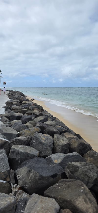 This is a full, vertical, outdoor, eye-level shot of a rocky shoreline in Kaaawa, Hawaii, United States of America. The foreground is dominated by a pile of large, dark gray and black rocks, some of which are partially submerged in the ocean. A light gray, concrete pathway runs along the top of the rocks on the left side of the image. A person in a white shirt and dark pants is walking on the pathway away from the camera. Further down the pathway, a stop sign with a stick figure walking is visible. The ocean stretches across the midground and background, with gentle waves lapping at a sandy beach on the right side of the image. Several people are visible swimming and wading in the ocean. The sky is overcast with thick, gray clouds, interspersed with patches of blue sky. A palm tree is partially visible on the far left.