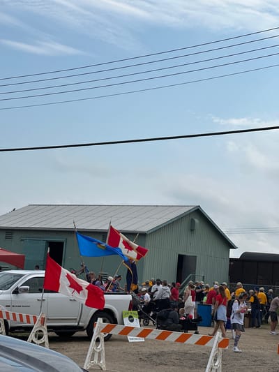 The image depicts an outdoor scene with a group of people gathered, many appearing to be spectators. A white pickup truck, adorned with Canadian flags, is the central object. Several people are standing or sitting in and around the truck. Some are holding flags and appear to be part of a procession or event. The truck has "RAM 1500" visible on its side.
In the background, a light-colored building with a sloped roof is visible, and further back, a dark train car or structure. The ground appears to be dirt or gravel. Orange and white safety barriers are placed near the truck. Overhead, power lines stretch across the frame against a bright, cloudy sky. The weather appears to be overcast or partly cloudy.
The presence of Canadian flags suggests a Canadian event, and the "City of Leduc, Canada" context places the scene specifically in Leduc. The people's demeanor is difficult to ascertain fully due to the image orientation and distance, but the overall atmosphere seems to be one of a community gathering or celebration. There is text visible on a sign attached to the truck that reads "EVENT ON NOW" and "DOWNTOWNLEDUC.CA". The overall impression is of a parade or festival.