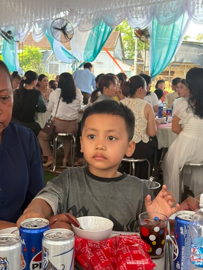 A horizontal medium shot depicts a young boy holding a clear polka-dotted glass, a white bowl, and several beer cans, seated at a table amid a gathering of people in what appears to be Bình An, Vietnam. The boy, with dark hair and light skin, looks directly at the camera. The table is covered with red snack packages and blue soda cans.

The background shows a crowded outdoor event with many people sitting at tables. The tables are decorated with turquoise and white draping, and there are fans attached to the structure above. The environment suggests a celebratory occasion, possibly a wedding or a large family gathering. The location is indicated as being in Bình An, Vietnam.