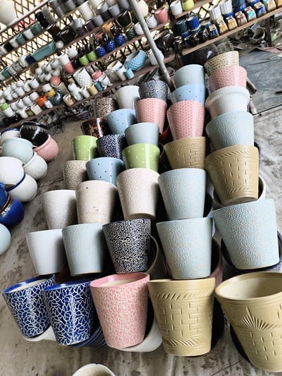 The image shows a large collection of ceramic flower pots of varying sizes, colors, and designs. They are arranged in stacks on the ground, and on shelves behind them. The pots on the ground seem to be larger than those on the shelves. The pots are various colors, including blue, white, pink, green, and beige. Some have patterns such as dots or textured designs, while others are plain. The location is Ghaziabad, India.