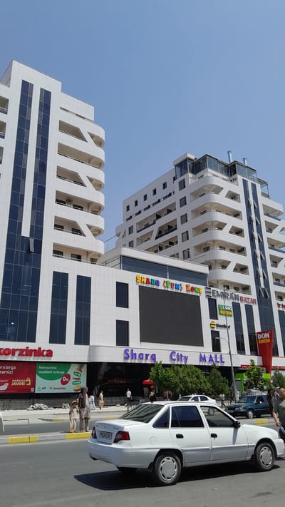 The image shows the "Sharq City Mall" in Denov, Uzbekistan. Two modern, white buildings with multiple balconies and dark-tinted windows dominate the scene. The building on the left is taller and features a distinct architectural design with curved balconies. The building on the right is also tall and modern, with a glass-enclosed rooftop section. A large LED screen is visible on the lower facade of the right building, and colorful signage reads "SHARQ FUN ZONE". Other signs include "EMIRAN BAZAR", "SHARQ BUUGER", "BeeFind" and "DON ARCHI". The mall appears to be a commercial hub.
In the foreground, a white sedan with license plate "S541 OA" is parked on the street. The street has a yellow and grey curb. To the left of the car, on the sidewalk in front of the mall, two people, likely a child and an adult, are walking. The child is wearing a straw hat and a light-colored outfit, while the adult is wearing a headscarf and similar-colored clothing. Several other pedestrians are visible further down the sidewalk near the mall entrance. Another car, a dark green sedan, is visible driving on the street behind the white car. Trees and other greenery are present behind the mall, suggesting an urban environment. The sky is clear and blue, indicating a sunny day. The overall impression is that of a bustling commercial area in Denov, Uzbekistan.