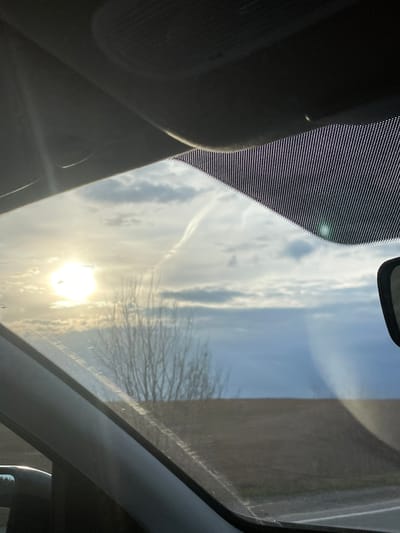 The image presents a view looking upwards through the panoramic roof or windshield of a vehicle, capturing a dynamic sky with the sun prominently featured. The perspective is from inside the car, directed towards the sky and reflections on the glass.
The sky above is a blend of clear blue patches and scattered white clouds. A brilliant, low-lying sun creates a strong glare and lens flare in the lower part of the frame. A faint, white contrail from an aircraft is visible cutting across the cloudy sky. Through the glass, the intricate, bare branches of a tree, indicative of winter or early spring, are discernible on the right side, partially masked by the sun's intensity and reflections. Further to the right, a dark, solid structure, possibly a building or a wall, is seen through the glass.
Elements of the car's interior frame the scene. On the left, a dark, textured surface, likely a part of the headliner or an interior trim, is visible. The edges of the glass, particularly the top-left and left sides, display a distinctive black frit band pattern, which consists of small dots. Minor streaks and smudges are present on the glass surface.
The low angle of the sun suggests the time of day is either early morning or late afternoon. The scene conveys a sense of being in transit or stationary within a vehicle, under a sunny and partly cloudy sky. The described location is Povarenki, Russia. No discernible text is visible within the image.
