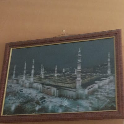 The image depicts a framed picture hanging on a plain light brown wall. The picture, enclosed within a rectangular dark brown frame with a gold inner border, displays an aerial or elevated view of a large, white-colored mosque complex. Numerous tall, slender minarets, at least eight distinct ones, are visible surrounding the main structure, which features extensive flat roof areas. The sky in the picture is dark, suggesting night or dusk conditions. The architecture is consistent with Al-Masjid an-Nabawi in Medina. The frame is suspended from a single small white hook or nail by a wire or string attached to its upper rear. No individuals or specific actions are present within the captured scene. This interior setting is located in the city of Pasir Muncang, Indonesia.