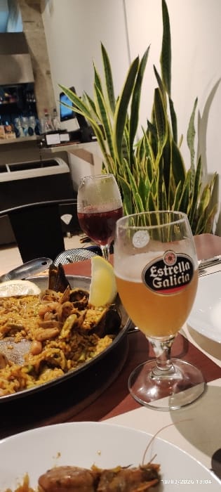 A dining table displays a feast. Beer in a glass with the "Estrella Galicia" logo sits near a glass of red wine and a slice of lemon. A black pan holds paella with visible mussels. Next to it, a white plate presents prawns. A decorative plant adorns the left side. The overall impression is a colorful and appetizing meal.