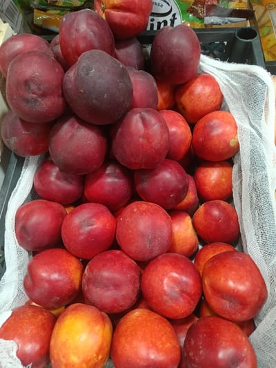 In Gulbakhor, Uzbekistan, a close-up, overhead view captures a black plastic crate overflowing with a vibrant display of ripe nectarines. The crate is lined with a white mesh fabric, adding a textured contrast to the smooth skin of the fruit. The nectarines are predominantly a deep red, with some exhibiting streaks of orange and yellow, indicating their ripeness and variety. They are packed tightly together, with some slightly piled on top of others, creating a sense of abundance. The lighting is natural and diffused, highlighting the glossy sheen of the fruit. In the background, slightly out of focus, are glimpses of colorful packaging, suggesting that this scene might be in a market or grocery store setting. The image is static, focusing solely on the arrangement of the fruit. There are no people or visible actions taking place.