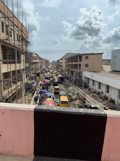 An elevated view captures a bustling street scene in Ebute-Metta, Nigeria, under an overcast sky. In the foreground, a concrete barrier, half painted pink and half black with a white stripe, frames the bottom of the image.
The street below, appearing unpaved and irregular with visible puddles and debris, is densely populated with people and vehicles. On the left, a multi-story building is under active construction, encased in extensive metal scaffolding. Beneath this structure, numerous market stalls operate, identified by varied tarpaulins and large umbrellas, with pedestrians moving between them and the roadway.
The roadway itself is occupied by multiple yellow commercial minibuses (danfo), dark-colored cars, and at least one white truck, some appearing stationary, others in motion. Concrete drainage elements or barriers are visible along and within the street, indicating ongoing infrastructure work or disarray.
On the right side of the street, a light-brown, multi-story residential building with multiple balconies stands, followed by a modern, flat-roofed white building. The area adjacent to these buildings along the street is cluttered with construction materials, debris, and what appear to be makeshift vendor setups or storage areas. Several individuals are visible in these areas, some sitting. A partially stripped vehicle, possibly a sedan, is observed near the white building.
The street recedes into the distance, maintaining its congested character with a continuous presence of vehicles and people, extending to the horizon where additional structures are vaguely visible.