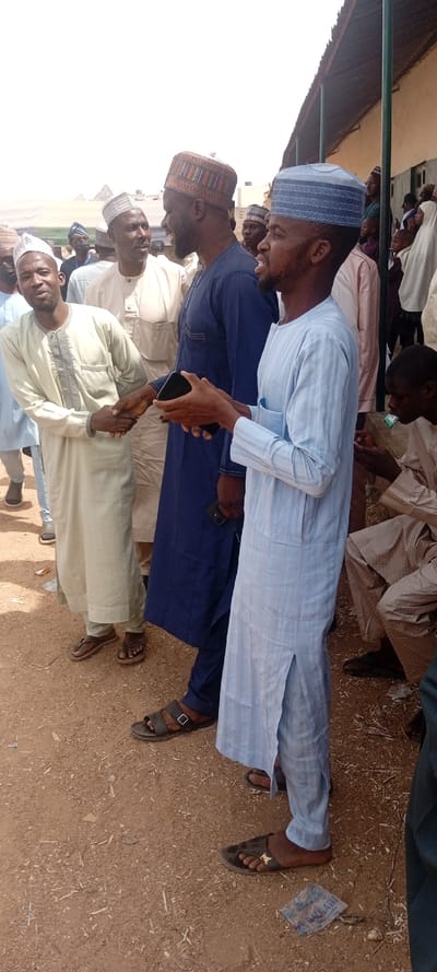 An outdoor scene in Sani Mai Nagge, Nigeria, depicts a daytime gathering of numerous individuals. In the foreground, from left to right, three adult males are prominent. A man in a light cream-colored long-sleeved tunic and matching pants, wearing a light-patterned cap, is engaged in a handshake with a man to his right. This second man is dressed in a dark blue long-sleeved tunic and matching pants, wearing a dark-patterned cap, and displays a slight smile. A black smartphone is visible in his left waist pocket or clipped to his attire. To his right, a third man, wearing a light blue striped long-sleeved tunic, matching pants, and a light blue patterned cap, holds a black smartphone horizontally with both hands, looking towards the left side of the frame. All three men wear open-toed sandals.
In the midground and background, a crowd of other individuals, primarily men in similar traditional attire, are visible standing. Some women in head coverings and several children are also present in the background near a structure. The ground beneath the individuals is unpaved, composed of reddish-brown dirt with scattered small debris and a discarded plastic wrapper visible in the lower right foreground. The background features a building with a dark, possibly corrugated, roof overhang supported by green vertical poles, and light-colored walls. The scene is illuminated by bright daylight, suggesting clear weather conditions. In the far lower right, a person is partially visible, seated, and appears to be consuming an item from a small white and green packet.