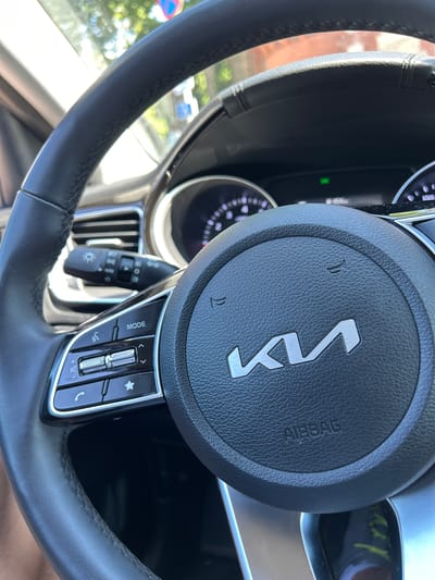 This media file is a close-up, eye-level, indoor shot of a car's steering wheel, specifically the driver's side. The focus is on the steering wheel's center hub and a portion of the left side. The steering wheel is black and appears to be made of leather, with visible stitching. In the center of the hub is the modern, stylized "KIA" logo in a metallic finish. Above and below the logo are small horn symbols. To the right of the logo, the word "AIRBAG" is embossed vertically.
To the left of the steering wheel hub, a part of the car's instrument cluster is visible, showing illuminated circular gauges with white markings against a black background. A green indicator light is also visible on the dashboard. On the steering wheel's right spoke, there are control buttons. These include a "MODE" button, directional arrows, a star icon, a volume control rocker, and a phone receiver icon. A turn signal lever is visible on the left side of the steering column.
The background suggests an outdoor setting with natural light. Through the car's windshield, blurred green foliage and some signs of an urban environment are discernible, including what appears to be a red and blue traffic sign. The overall lighting suggests it is daytime, likely sunny. There are no people visible. The scene is a static capture of the car's interior. Visible text includes "KIA", "AIRBAG", and "MODE". The location is identified as Tartu, Estonia.