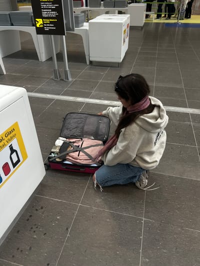 A young woman wearing a grey hoodie with "EST. YEAR OF" written on it, blue jeans, and a pink scarf, is kneeling on the grey tiled floor of the Granadilla, Spain, airport. She is facing a suitcase with the top open, partially revealing a pink backpack and other items. The suitcase is on the floor next to a white wall with a yellow sticker that reads "Glass / Vidrio". The background is a blurry view of the airport with some white furniture and white walls. The woman is preparing for travel, possibly at the baggage claim or security checkpoint.