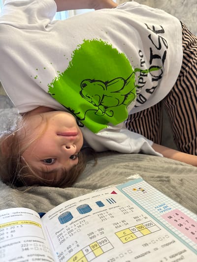 A child with shoulder-length brown hair is lying on a gray textured surface with an open math workbook. The workbook features columns of numbers and mathematical problems, some in Cyrillic script. The child is wearing a white long-sleeved shirt with a large green graphic and black text. The child is also wearing striped pants. The image shows the child looking directly into the camera. It is impossible to identify the city, Yaroslavl', based on visual information.