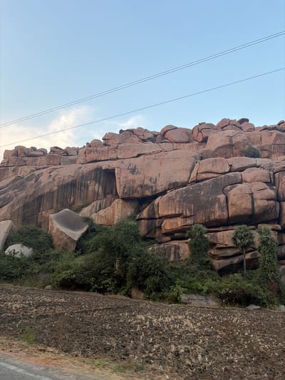 The image depicts a natural rock formation, likely a granite outcrop, situated in Chikkarampur, India. The formation is composed of numerous large, reddish-brown, rounded boulders stacked irregularly, creating a steep, stratified appearance. Dark vertical streaks are visible on some rock surfaces, indicative of erosion or water runoff. On the lower left slope of the formation, a large, lighter gray, somewhat cuboid boulder with a concave upper surface rests amidst dense green shrubbery. Below this, a field of dark, tilled earth with scattered dry plant stubble occupies the foreground. Several tall, slender palm trees are partially visible on the right side of the rock formation, integrated with the surrounding green vegetation. The sky above is light blue with sparse, wispy white clouds. Two parallel utility wires traverse horizontally across the upper portion of the sky. No human subjects, animals, or direct actions are depicted. The perspective appears to be from ground level, possibly adjacent to a road, evidenced by a faint orange line at the bottom edge.