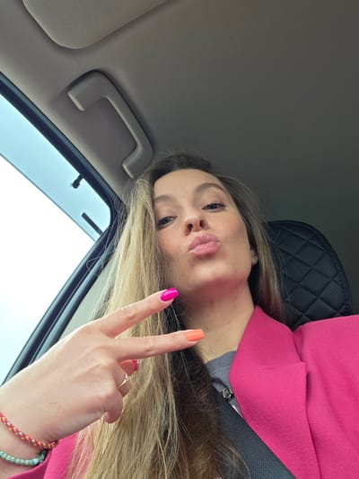 A woman is inside a car. She is wearing a pink blazer and has on a seatbelt. She is holding up her hand in a peace sign. Her nails are painted bright pink and orange. The inside of the car is gray. The location in the city of Аур, Russia, is unknown.