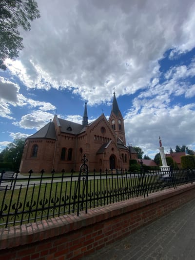 A red brick church with a tall, dark spire stands against a backdrop of a blue sky filled with dramatic white clouds. The church has arched windows and a clock face on its tower. In front of the church is a black metal fence, and beyond that, a white pedestal with a statue of a figure in red and white clothing. To the right of the pedestal, another smaller statue stands atop a green hedge. The scene is located in Opatów, Poland.