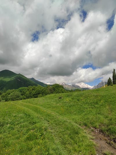 This is a landscape photograph taken in the city of Kumlistsikhe, Georgia. The image depicts a verdant, rolling green meadow in the foreground, dotted with small yellow wildflowers. A dirt path is visible, winding through the grass. In the mid-ground, a dense line of lush green trees forms a natural border. Beyond the trees, a mountain range rises, with the lower slopes covered in green vegetation and the peaks dusted with snow. The sky above is a dramatic mix of dark, brooding clouds and patches of bright blue sky. Power lines and utility poles are visible on the right side of the frame, with tall, slender trees standing nearby. The overall impression is of a natural, mountainous landscape under a dynamic sky.