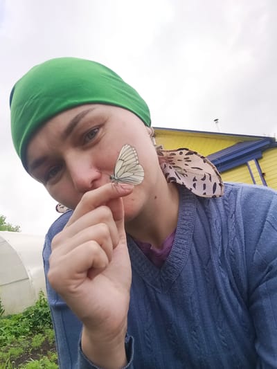 An adult individual is depicted in an outdoor low-angle portrait in Gabdyukovo, Russia, under an overcast sky. The person has light skin, light-colored eyes, and wears a solid green head covering. They are dressed in a blue cable-knit V-neck sweater over a purple top. A small dark mole is visible on the neck below the left ear. A white butterfly with prominent dark veins is perched on the tip of the person's right index finger, which is held near their nose. A second butterfly, displaying light-colored wings with dark irregular spots on the underside, is positioned on the person's left shoulder near their ear. The background includes a white arched greenhouse and green garden plants to the left, and a building with bright yellow siding, a dark blue roofline, and a visible chimney to the right.