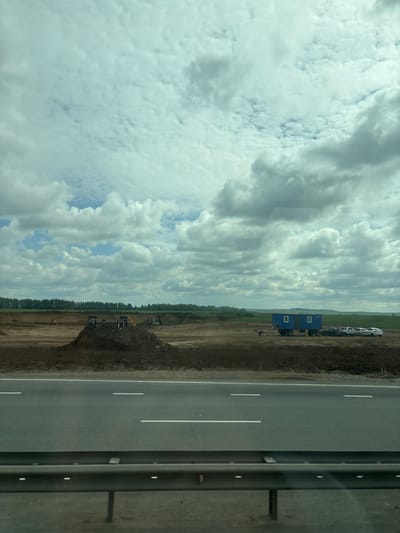 The image captures a construction site located in the city of Poisevo, Russia. The scene unfolds under a sky filled with dramatic, fluffy white clouds against a pale blue backdrop. In the foreground, a multi-lane asphalt road with white lane markings runs horizontally across the frame, separated by a metallic guardrail. Beyond the road, the ground is predominantly brown and barren, indicative of ongoing excavation and earthmoving. Several pieces of heavy machinery, including what appear to be excavators, are visible scattered across the site, some positioned near large mounds of dirt. To the right of the construction area, a blue portable trailer is parked, accompanied by a row of several white and black passenger vehicles. A single person can be seen sitting near the blue trailer. In the distance, a dense line of green trees forms a natural boundary, beyond which the terrain appears to be agricultural fields under the expansive cloudy sky.