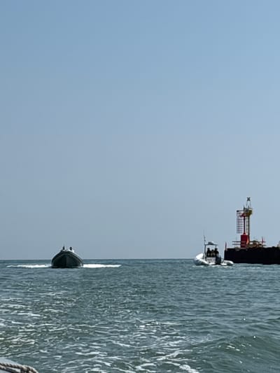 The image shows a body of water with two boats and a large structure in the background. The sky is clear and blue. The boat in the foreground on the left is dark-colored and appears to be moving, leaving a wake. Two people are visible in this boat. On the right side of the image, a lighter-colored boat with a canopy is carrying at least three people. Behind this boat is a large, dark, barge-like structure with a tall, red and yellow tower-like element on top, which appears to be a navigational marker or part of a construction or maintenance platform. The water is choppy with whitecaps. The scene is likely a marine environment, possibly the Adriatic Sea off the coast of Cesenatico, Italy, given the presence of marine structures.