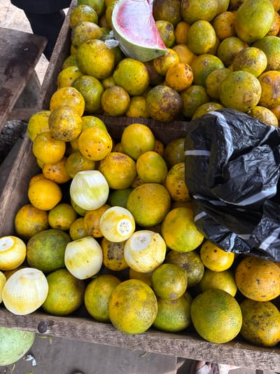 This image depicts a market stall in Ogere, Nigeria, overflowing with a large quantity of citrus fruits, primarily oranges. The oranges display various shades of yellow, green-yellow, and orange, many exhibiting natural blemishes and dark speckles. Several oranges appear partially peeled or cut, exposing white pith. A slice of watermelon, featuring pink flesh and dark seeds, wrapped in clear plastic, rests atop a pile of oranges near the upper center. A crumpled black plastic bag with white horizontal stripes is situated on the right side, partially covering some fruits. The fruits are contained within a rustic, weathered wooden structure, likely a crate or display stand. In the upper left corner, a dark-clothed individual is partially visible, while at the bottom, light-colored footwear suggests the presence of another person near the stall. A large, green-skinned fruit, possibly a watermelon, is partially visible at the bottom left.
