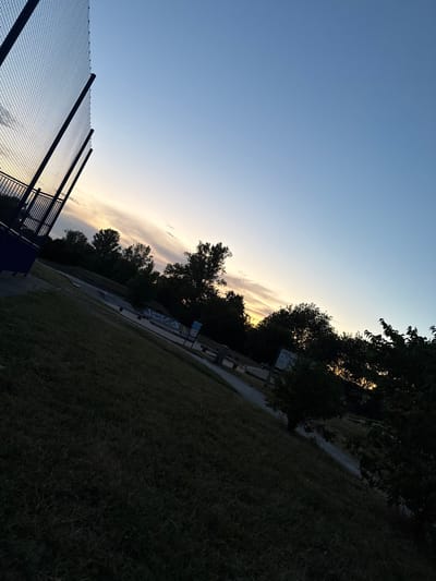 The image depicts a park area at dusk. A tall chain-link fence, possibly part of a sports facility, dominates the left side of the frame, angled upwards. The sky above transitions from a pale yellow and orange near the horizon to a deep blue higher up, indicating the setting sun. Below the fence, a grassy slope descends towards a park with trees and what appears to be a skate park in the middle ground. The grass in the foreground is dark and appears somewhat overgrown. Silhouetted trees line the background. There are no people or animals visible in the scene. The location is identified as Saint-Orens-de-Gameville, France.