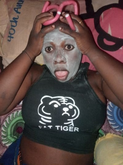 A dark-skinned woman wearing a black tank top that features a white cartoon drawing of a tiger with the words "FAT TIGER" printed below. She has a white facial mask and pink hair rollers in her hair. Her hands are placed on top of her head. The woman's mouth is open with her tongue sticking out. She appears to be lying on a bed or couch that has several different patterned pillows.