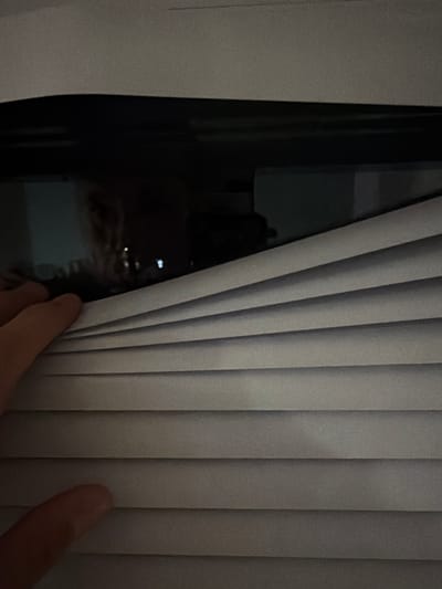 A person's hand is pulling open white blinds, revealing a dark interior with a blurred reflection of a person's face in what appears to be a window. The scene is indoors, likely in an apartment or house. There is no information available to determine the specific location in Sosnowiec, Poland.
