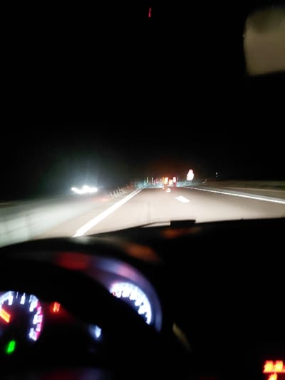 A nighttime car interior view, captured from the driver's perspective. The illuminated dashboard gauges glow in the darkness. Outside, blurry lights from other vehicles create streaks along the highway. The road stretches ahead, dimly lit. A dark steering wheel dominates the foreground.