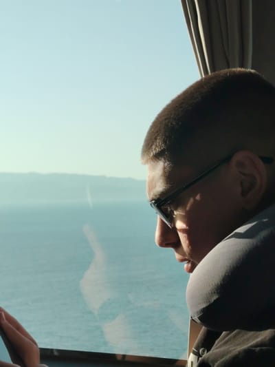 A close-up, side profile shot captures a person, likely male, looking contemplatively out of a window. The individual, positioned on the right side of the frame and facing left, has short, dark hair and wears dark-rimmed glasses. A grey travel neck pillow is snugly around their neck, suggesting comfort during travel or rest, and they are wearing a dark t-shirt.

Their gaze is directed towards the expansive outdoor scenery visible through the window. A vast body of calm, blue-green water dominates the midground, stretching towards a hazy horizon where distant, light-colored mountains or landmasses are faintly visible. The sky above is a clear, bright pale blue, indicating sunny weather during daytime, possibly morning or afternoon, given the direct sunlight illuminating the person's face and hair.

A hand is partially visible at the bottom left of the frame, suggesting the person might be holding something out of view. Inside the setting, a dark, pleated curtain or blind is visible in the upper right corner, indicating an enclosed space such as a vehicle (like a bus, train, or boat) or a room.

Considering the location context of Iznik, Turkey, the large body of water is consistent with Iznik Lake, and the distant landmasses could be its surrounding mountains or shores. The image evokes a tranquil moment of observation or travel by the lake. No discernible text is visible within the frame.