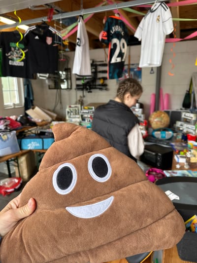The image depicts an indoor scene in Deinste, Germany, consistent with a garage sale or flea market setup. In the immediate foreground, a person's hand, partially visible on the left, holds a brown, plush, smiling poop emoji pillow. The pillow features white-rimmed black eyes and a white smile, rendered in embroidery. A white tag, partially obscured, is visible on the lower right side of the pillow, displaying "Desire P...".

In the middle ground, a female individual stands with her back to the camera, facing a table filled with various items. She has braided hair and wears a black puffer vest over a white long-sleeved shirt. She appears to be examining merchandise.

The background shows a cluttered utility or garage space. Soccer jerseys are prominently hung from a metal rail system across the ceiling. From left to right, these include a dark jersey with "BRASIL" and "CLAUDIO" text, a black Adidas jersey with white stripes and a crest, a dark patterned jersey with "LAVERZ 29", and a white Real Madrid Adidas jersey. Colorful streamers in yellow, pink, green, and orange are draped around the ceiling area.

To the left, a window provides natural light, and below it, a wooden table is laden with piled items and blue storage containers. To the right, the table where the female is standing is densely packed with miscellaneous objects, including a small globe, a blue portable stereo system, a black mini-oven or toaster, and numerous smaller items such as toys, books, and household goods. A pink hula hoop or similar object leans against the wall in the far right background. The walls are light-colored and appear to be unadorned.