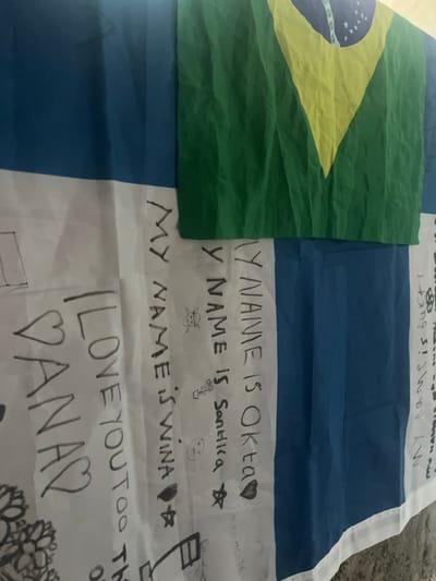 A fabric banner, located in Desa Batu Layar, Indonesia, is depicted vertically. The banner consists of a smaller, creased section of a Brazilian flag (green background, yellow rhombus, blue circle with white stars) overlaid on the upper portion of a larger, striped fabric. The larger fabric features alternating vertical blue and white stripes. The white stripes are extensively covered with handwritten black marker inscriptions and drawings. Legible text includes "I LOVE YOU TOO TH", "♡ DANAS", "MY NAME IS OKTA ♡", "MY NAME IS Santika ☆", and "MY NAME IS WINA ♡ ☆". Drawings observed include hearts, stars, a skull and crossbones, a small cartoon figure, and floral designs. Additional illegible text and drawings are present on the rightmost white stripe. No individuals are present.