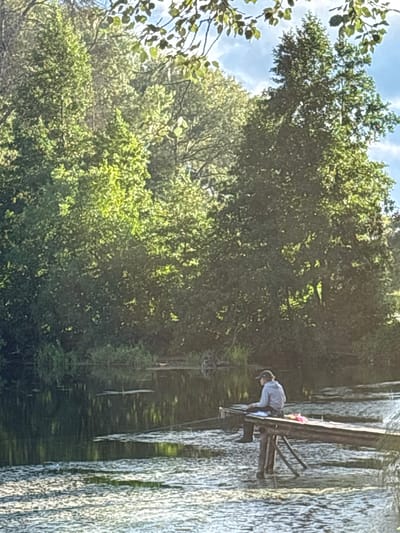 A person sits on a wooden pier extending into a body of water, likely a river or lake, surrounded by lush green trees. The person is wearing a baseball cap, a light-colored hooded sweatshirt, and dark pants. They are facing away from the camera, looking out at the water and appears to be reading or writing on a flat object. A fishing net is placed beside them on the pier, along with a pink object that may be a piece of clothing or a towel. The water's surface is dappled with sunlight and reflects the surrounding vegetation. The background is dominated by dense foliage, with some branches and leaves visible in the foreground against a blue sky with scattered clouds. The scene suggests a serene, natural setting, possibly in a rural or park area within Муромцево, Russia, given the context.